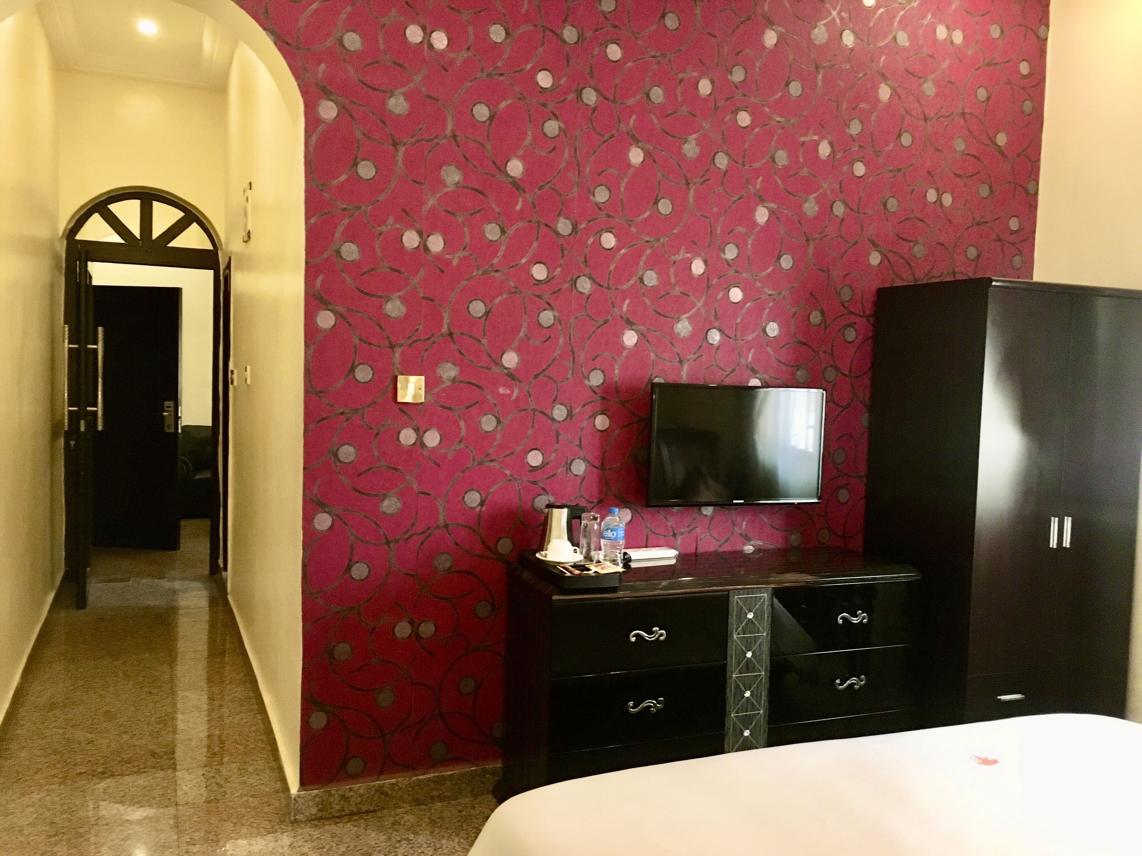 A hotel room with a red patterned wall, a flat-screen TV mounted above a dark wooden dresser, and a bed with white bedding.