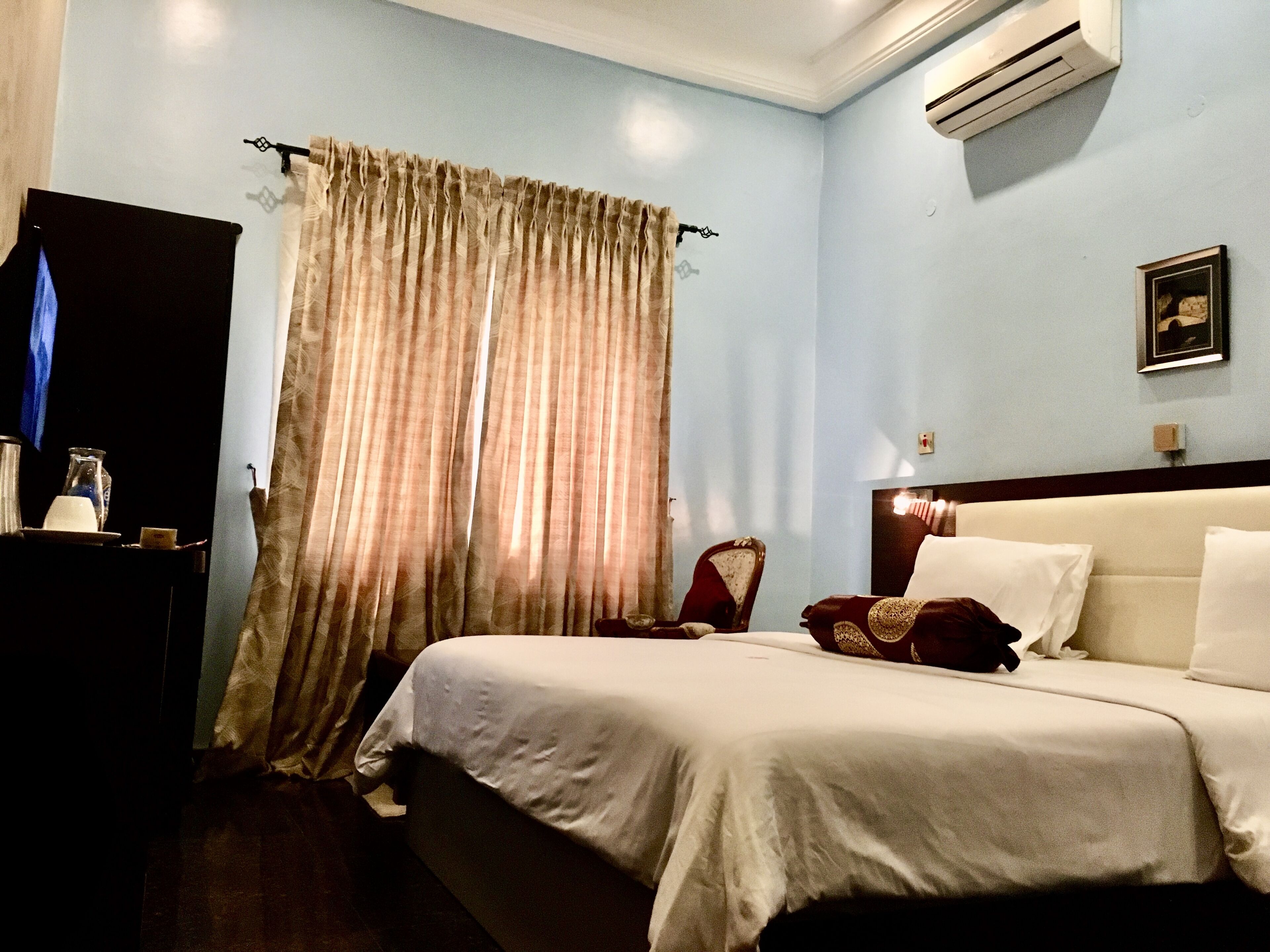 A hotel room with a bed, a chair, a TV, and an air conditioning unit.