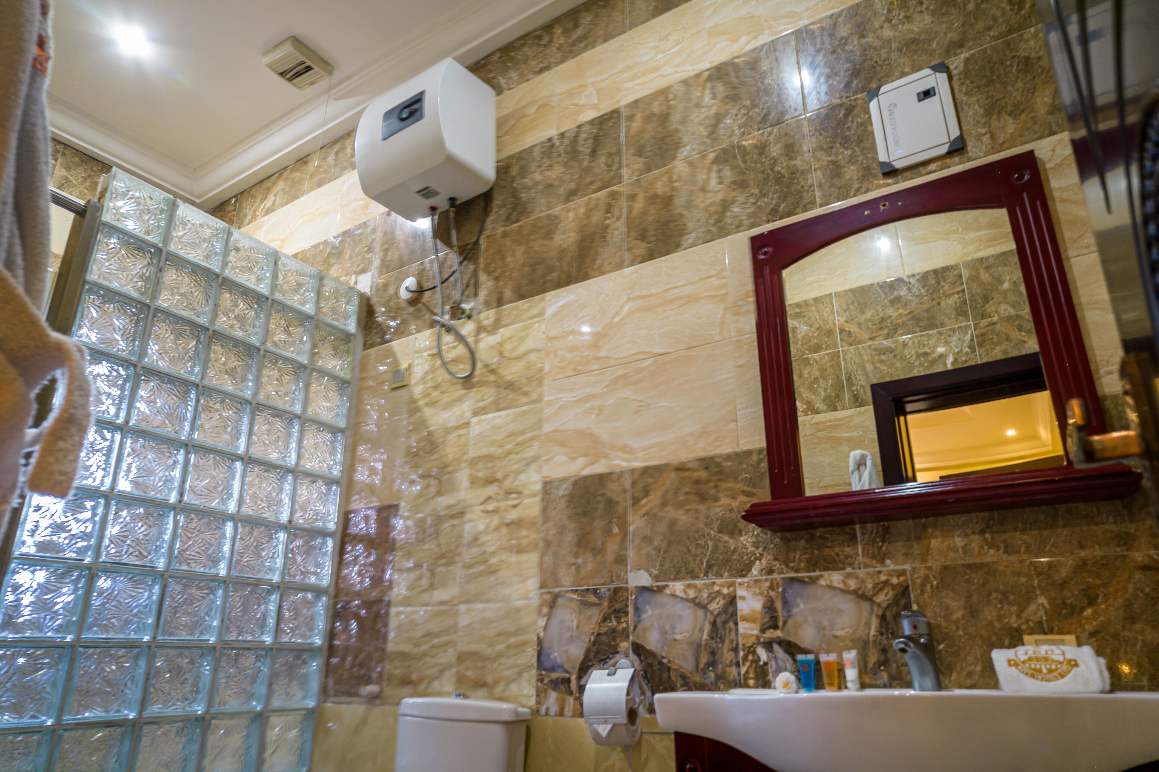 A bathroom with a marble wall, a glass shower enclosure, a sink with toiletries, and a mirror with a red frame.