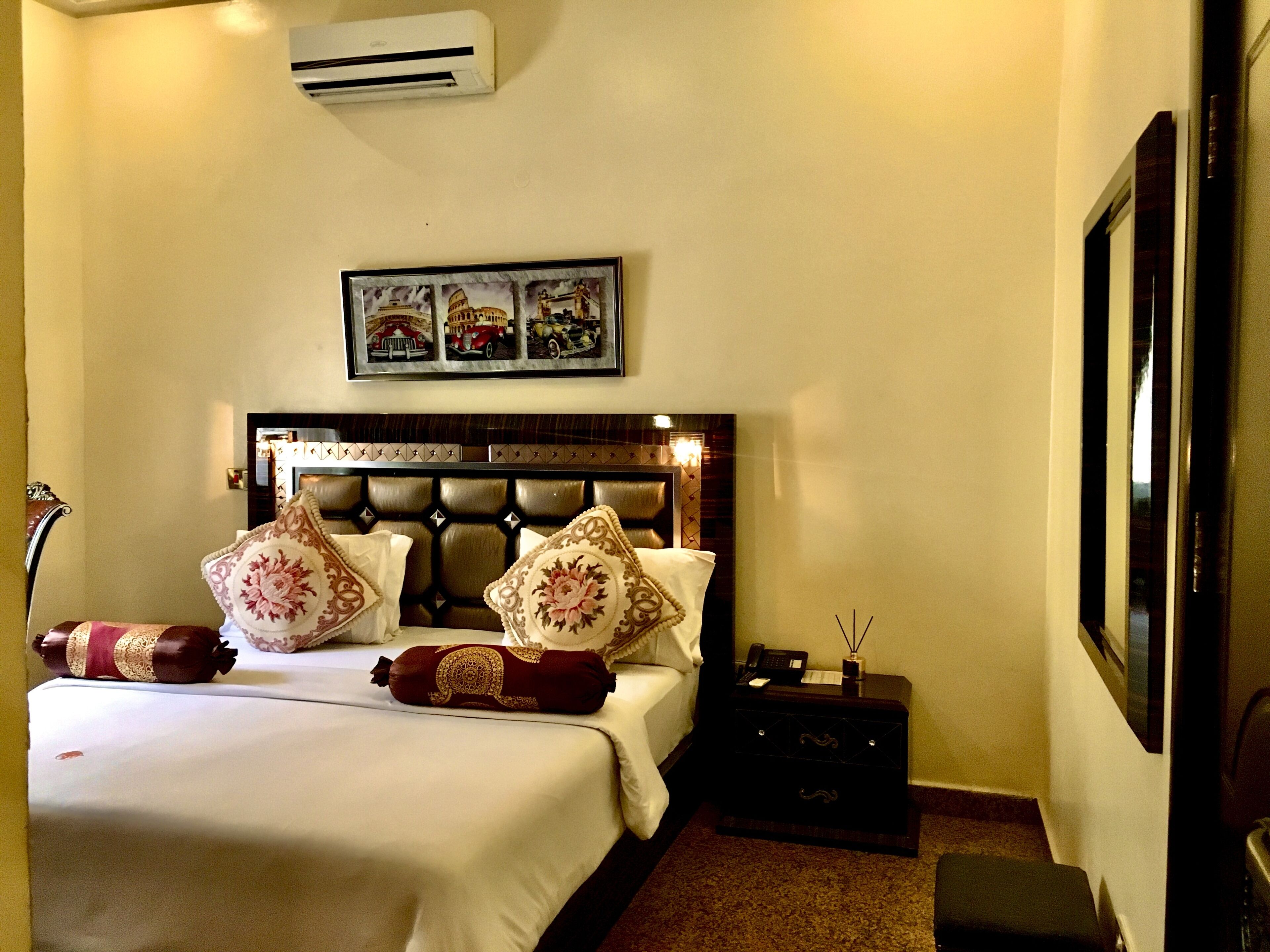 A hotel room with a large bed, a headboard with a decorative panel, a nightstand with a lamp, and a framed picture above the bed.