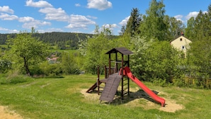 A playground with a slide and climbing structure in a grassy field with trees and a house in the background.