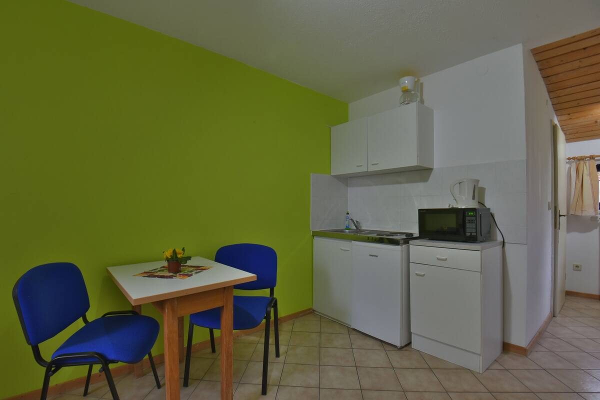 Een kleine keuken met een magnetron, gootsteen en kastjes. Er is een eettafel met twee stoelen.