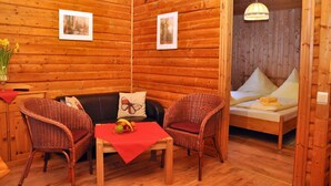 A cozy room with wooden walls, a sofa, two wicker chairs, a small table with a fruit basket, and a bed with pillows.