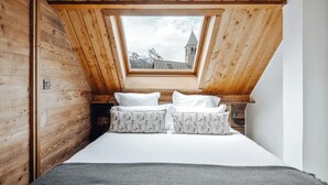 Een slaapkamer met schuine wanden, houten lambrisering, een bed met wit beddengoed en bloemenkussens, en uitzicht op een berg en een kerk door een dakraam.