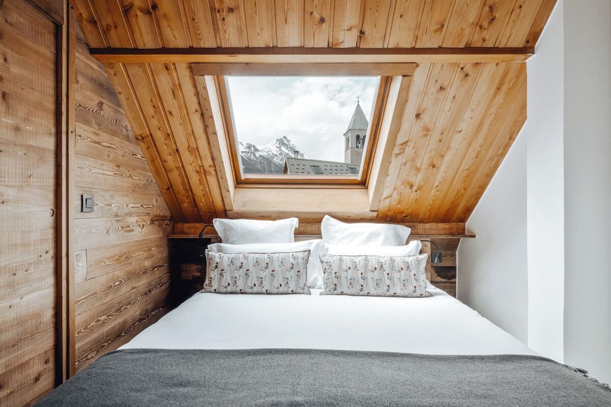 Ein Schlafzimmer mit schrägem Dach, Holzwänden, einem Bett mit weißer Bettwäsche und floralen Kissen sowie Blick auf einen Berg und eine Kirche durch ein Dachfenster.