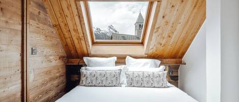 A bedroom with a sloped ceiling, wooden walls, a bed with white bedding and floral pillows, and a view of a mountain and a church through a skylight.