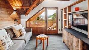 Een gezellige woonkamer met een houten plafond, een bank met kussens, een klein tafeltje met boeken, een tv op een houten dressoir en een schuifdeur naar een buitenruimte.