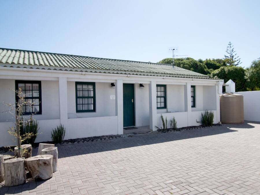 A white building with a green door and windows, surrounded by potted plants and a paved area.