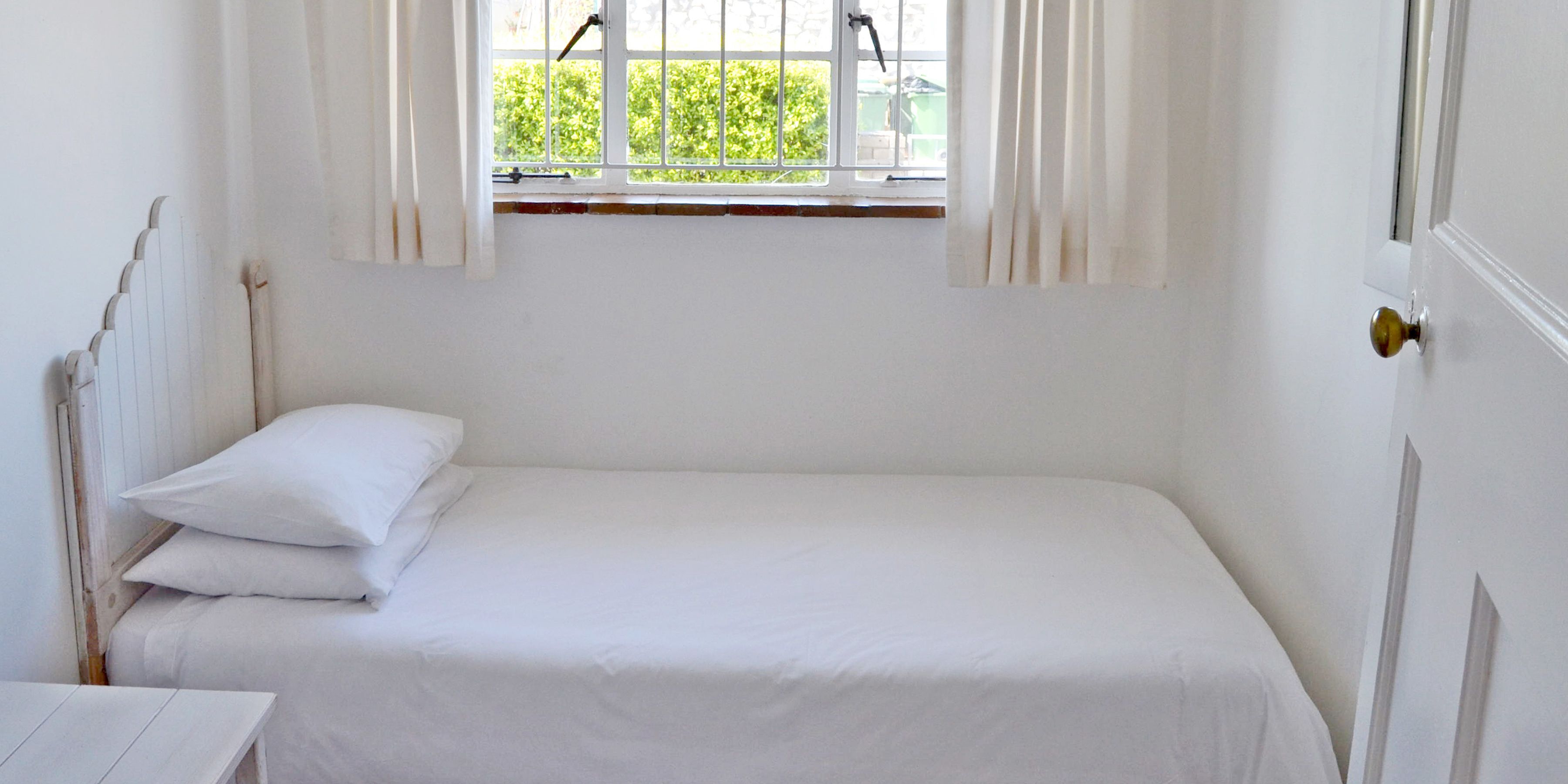 A small, white, single-bed room with a window and curtains.