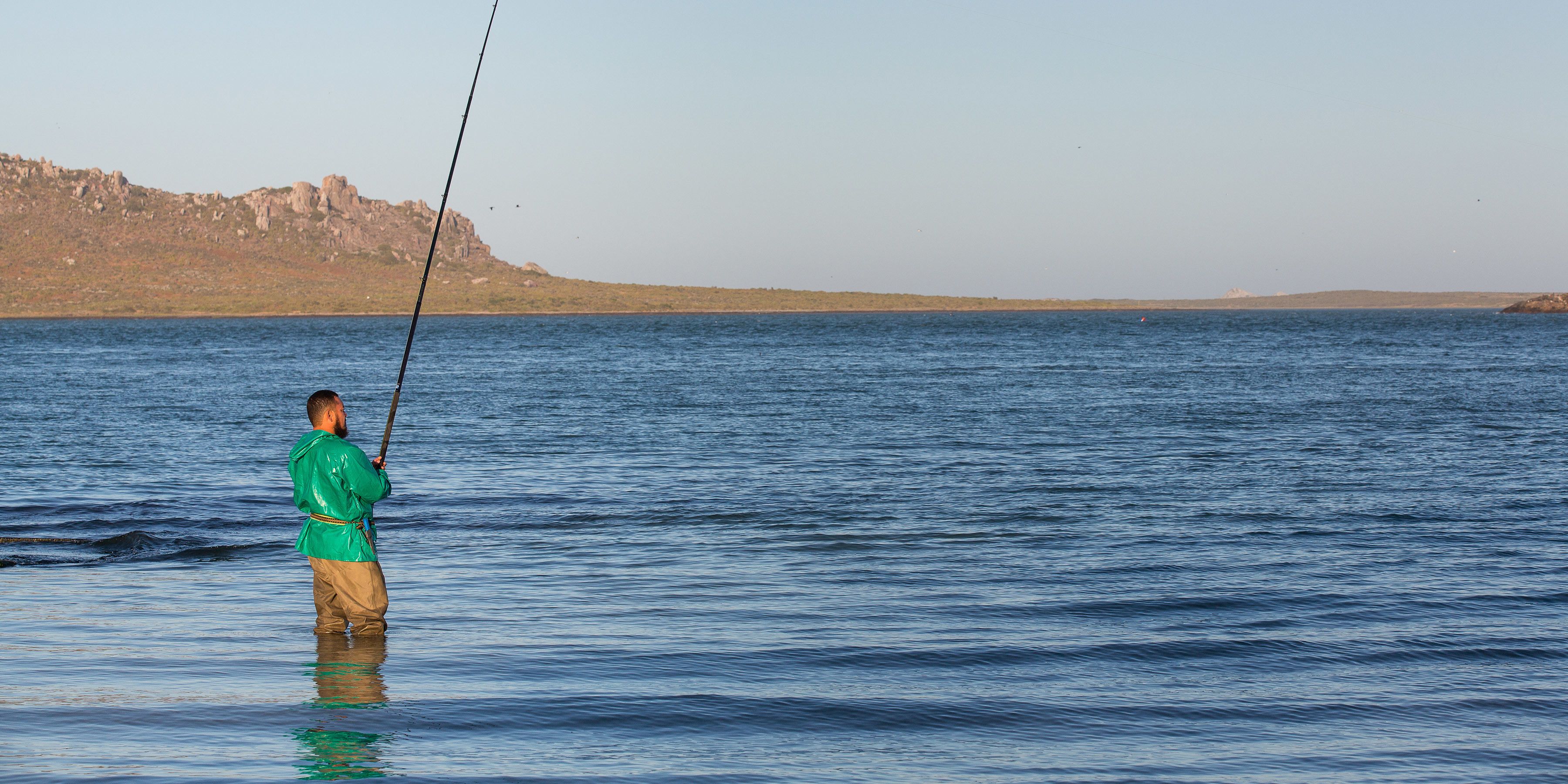 A person fishing in a calm body of water with a rocky landscape in the background.