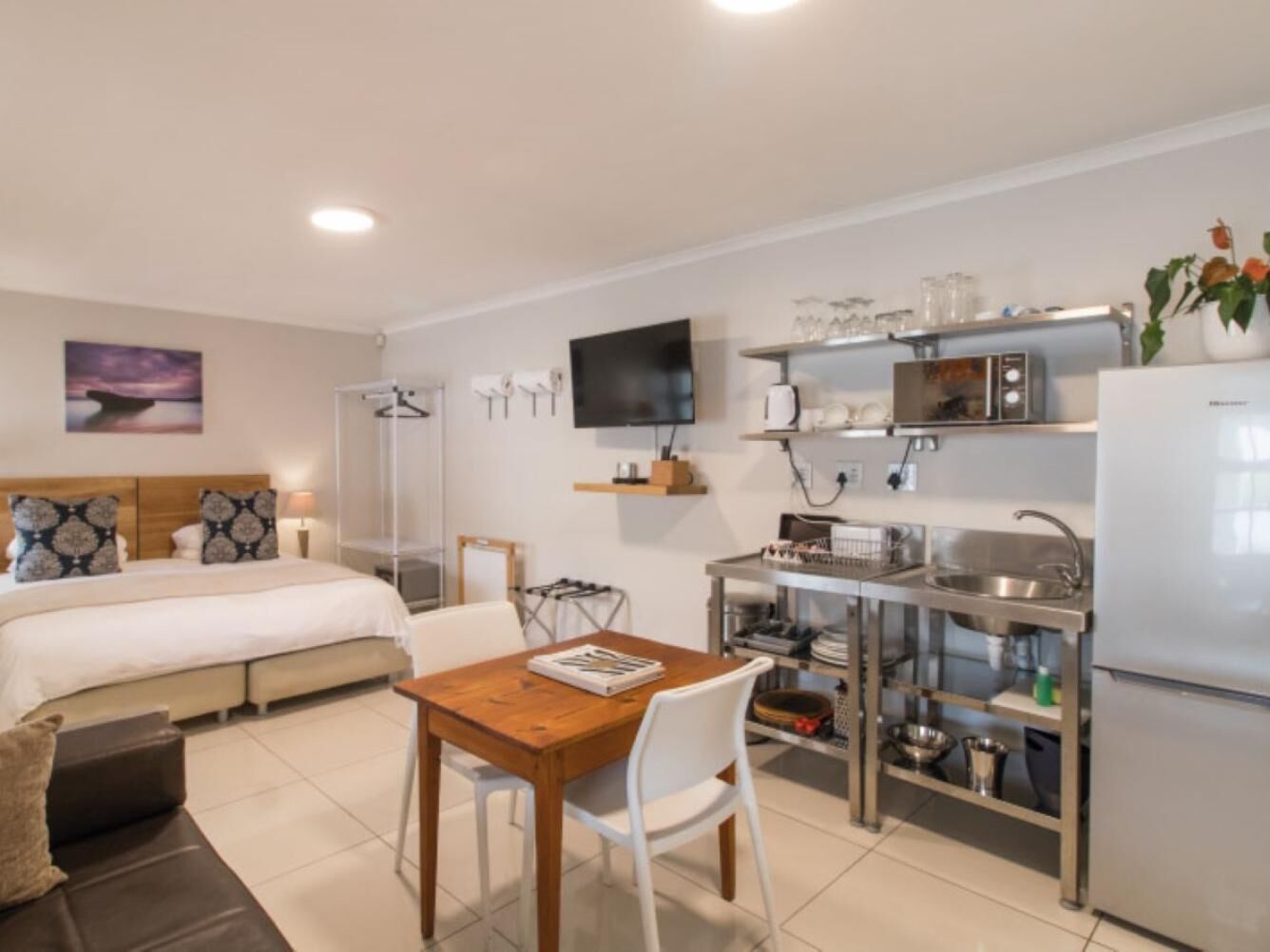 A modern hotel room with a bed, a dining table with chairs, a kitchenette with a microwave, and a TV mounted on the wall.