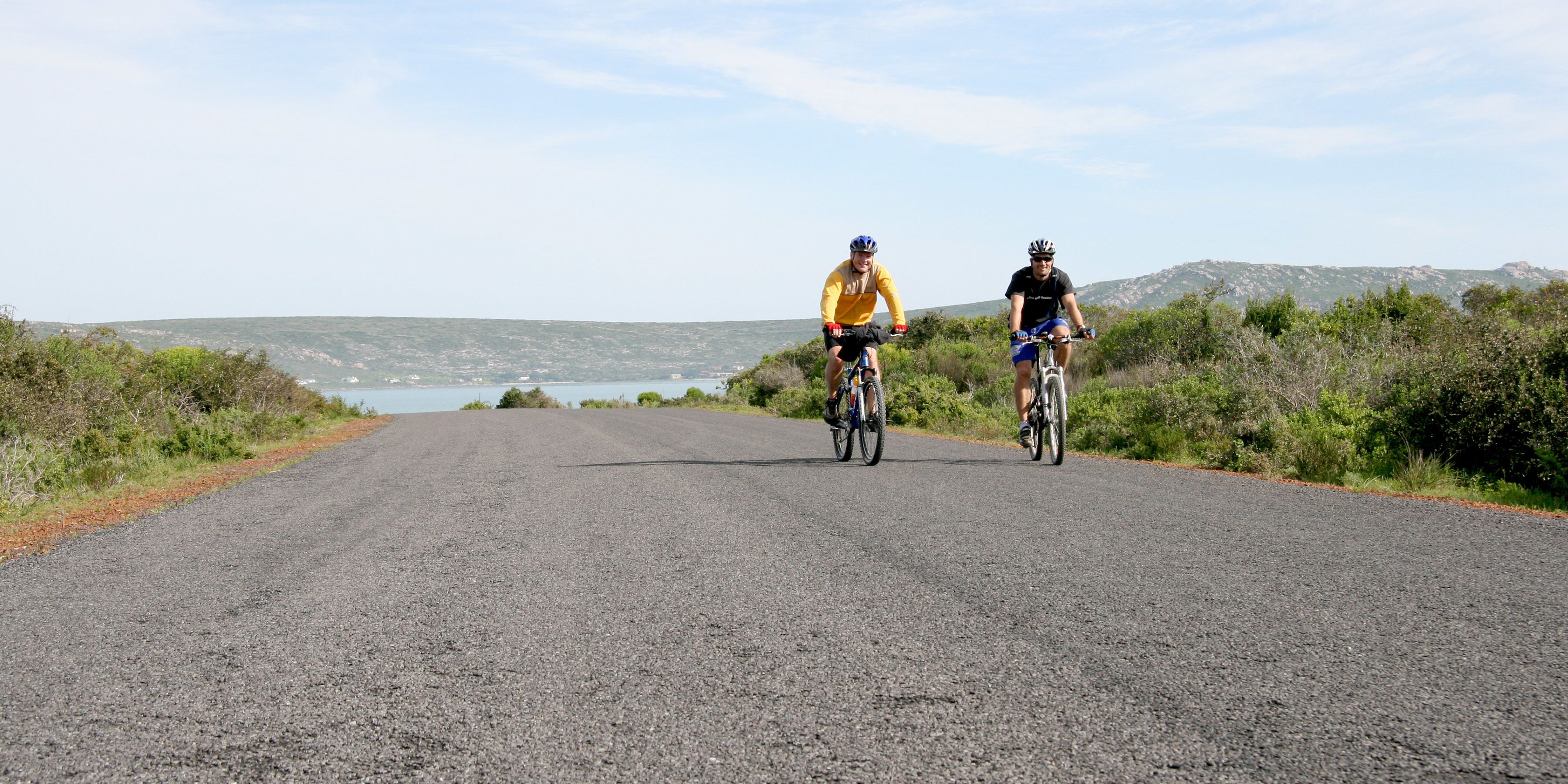 Deux cyclistes sur une route asphaltée, avec une vue panoramique sur des collines et un plan d'eau au loin.