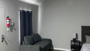 Ett modernt rum med en grå soffa, ett träbord, en säng med ett vitt överkast, ett nattduksbord, en takfläkt och en brandsläckare på väggen.