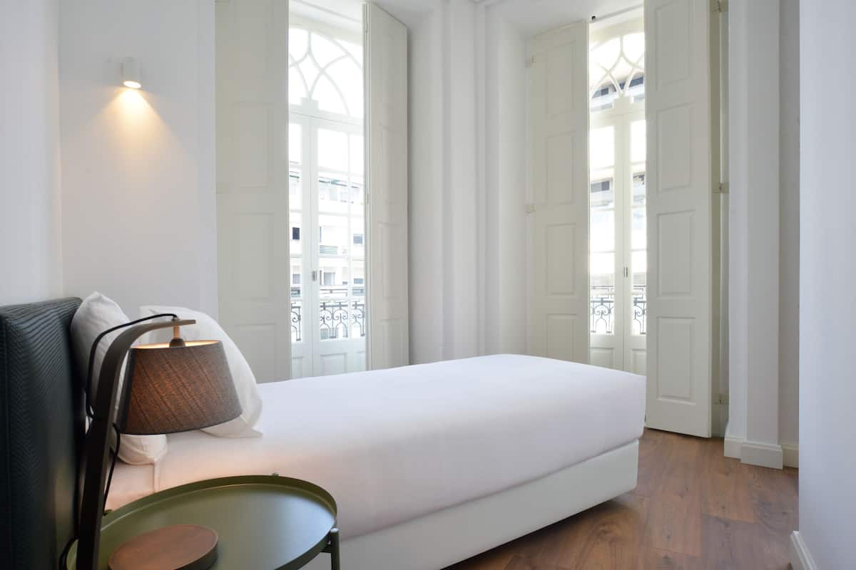 A minimalist bedroom with a white bed, a green side table, and a lamp.