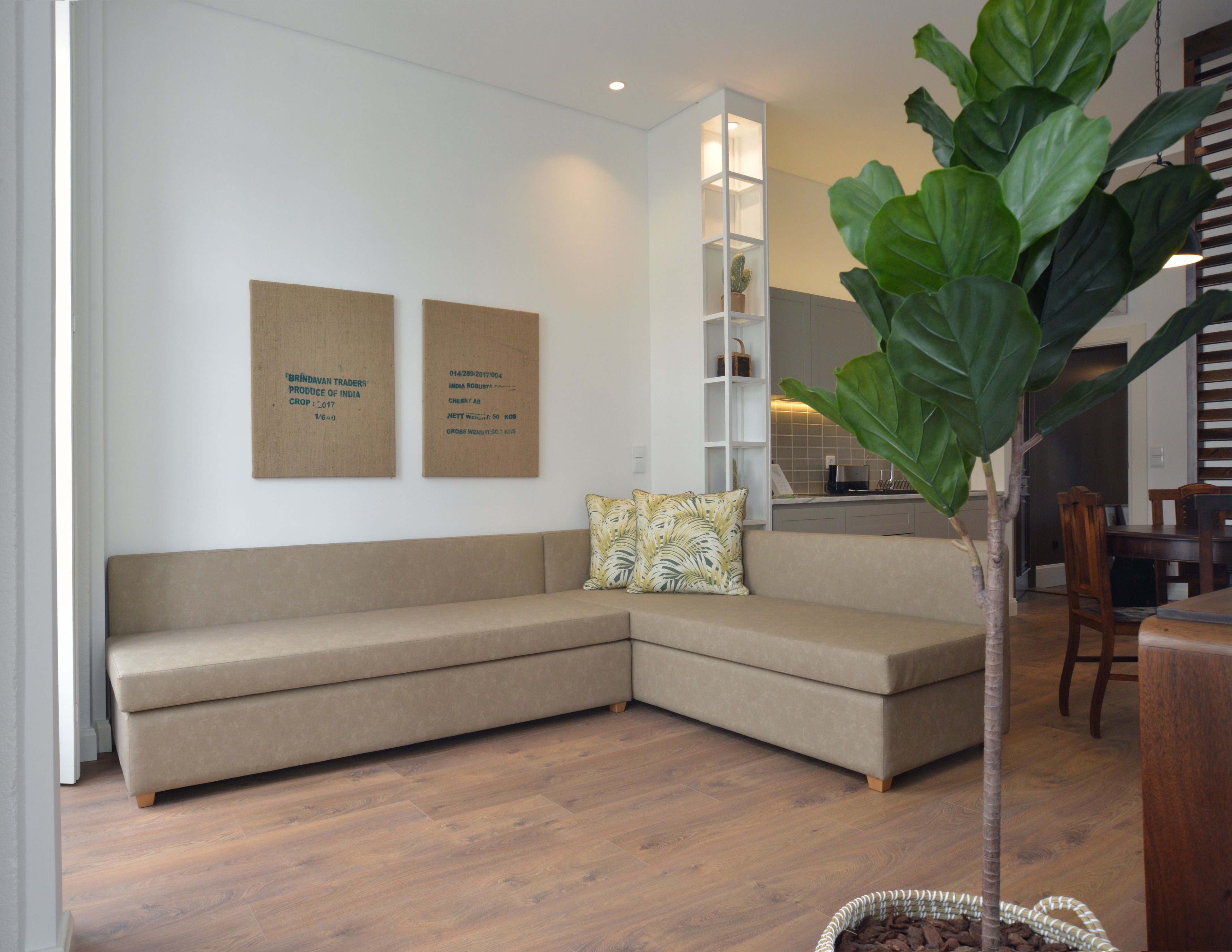 Una sala de estar moderna con un sofá seccional color beige, una mesita de centro pequeña y una planta verde grande. Hay dos diplomas enmarcados en la pared y una estantería blanca al fondo.