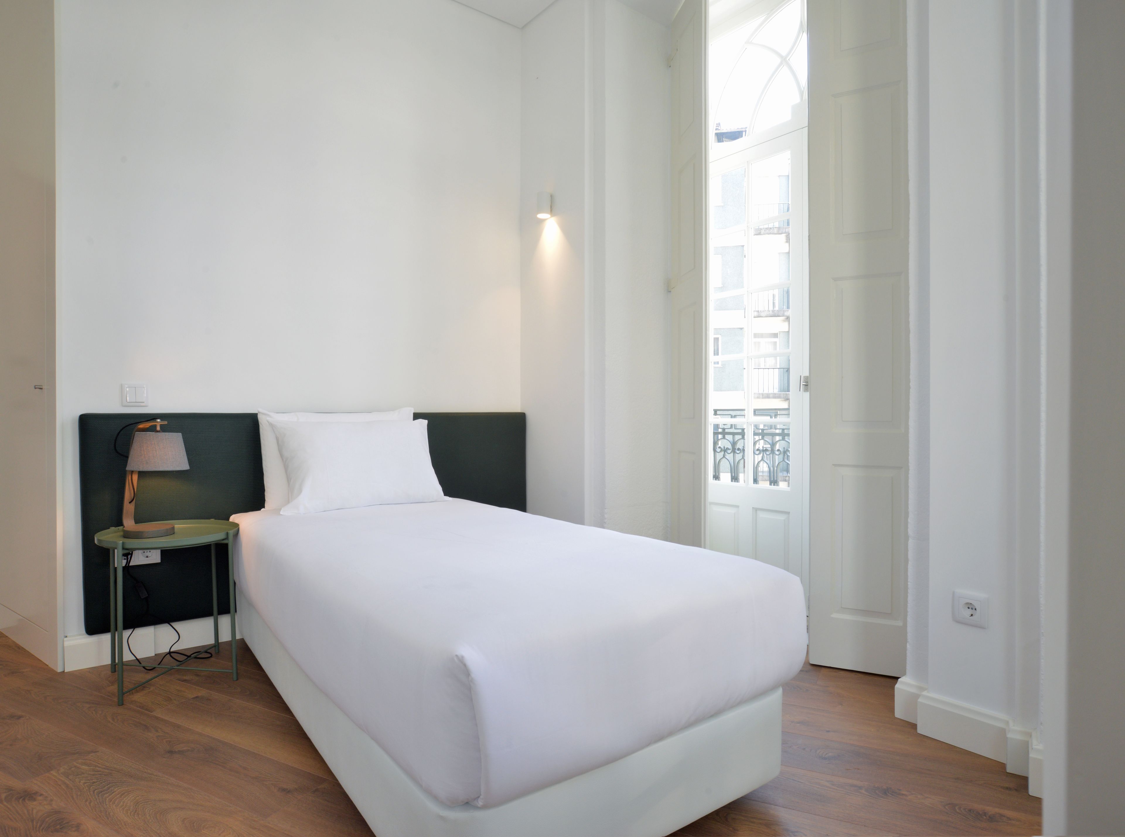 Un letto rifatto con cura in una stanza con un comodino, una lampada e vista sull'esterno tramite una finestra.