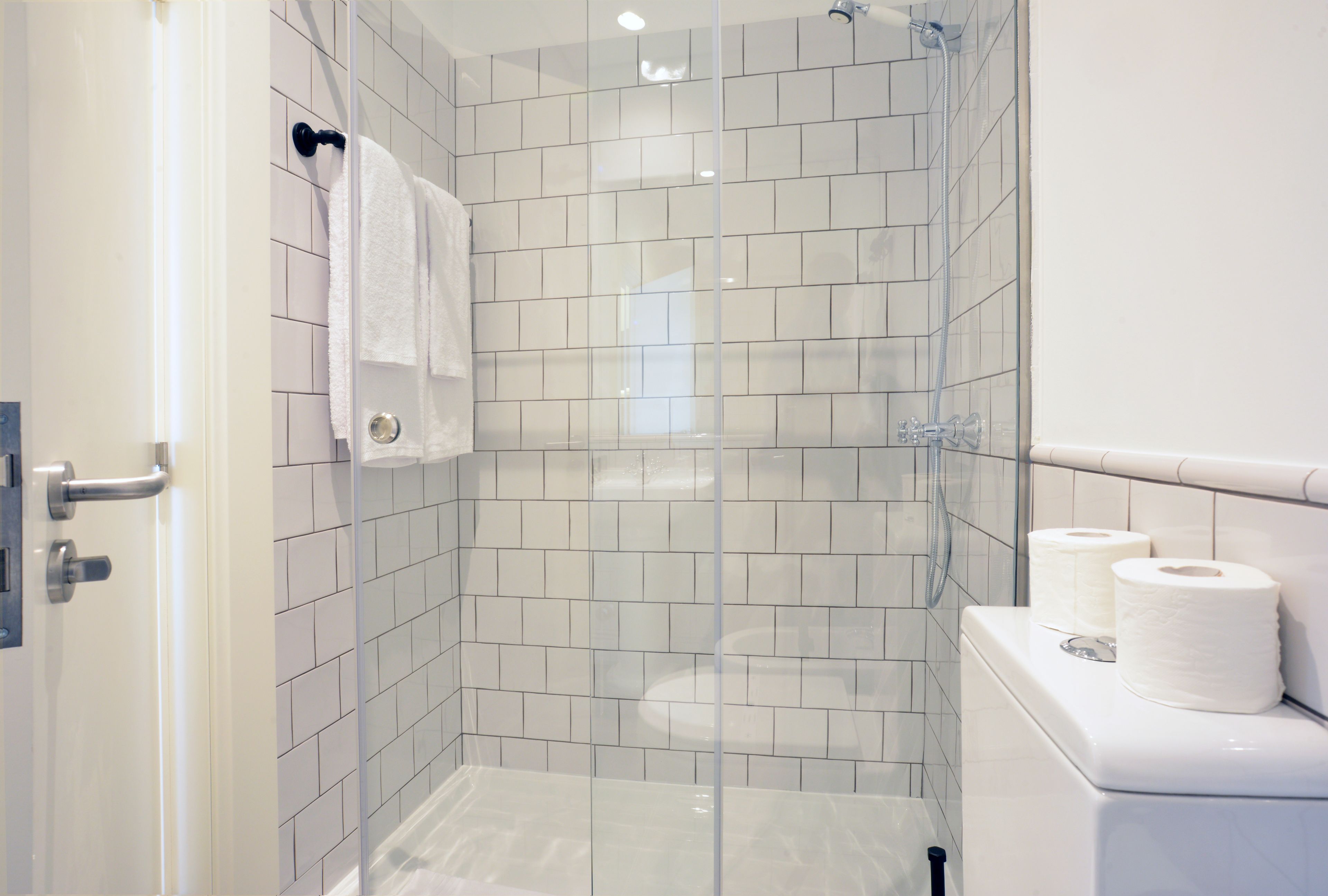 A modern bathroom with white tiles, a glass-enclosed shower, a toilet with toilet paper, and a towel rack.