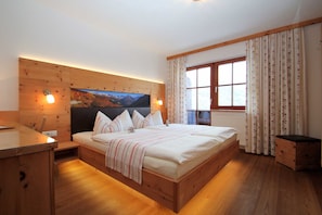 A wooden bed with a headboard, a bedside table, a window with curtains, and a wall-mounted TV.