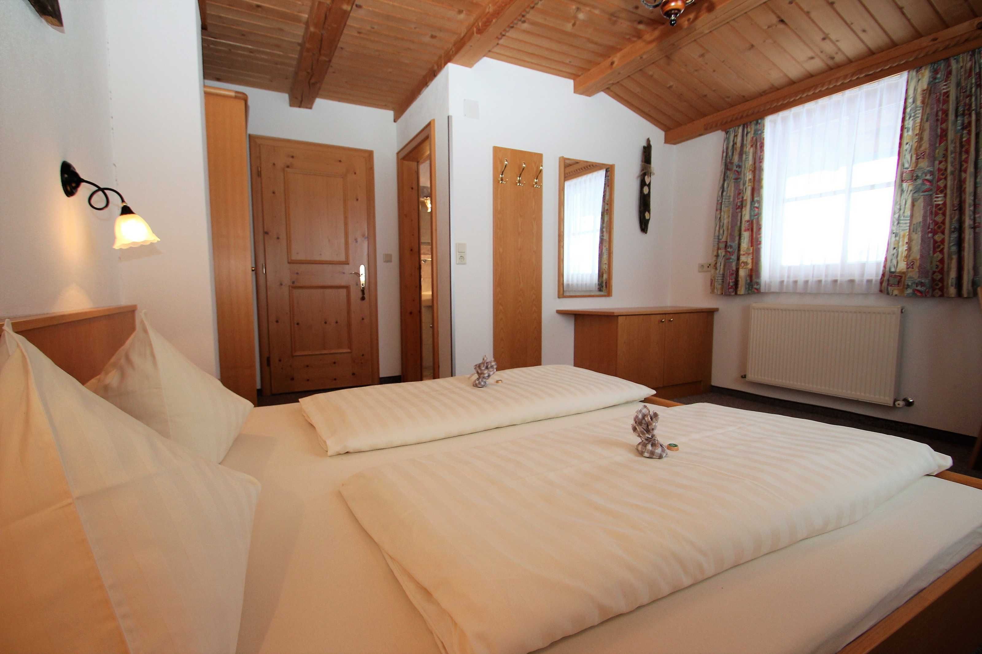 A room with two beds, wooden ceiling beams, a wall-mounted light fixture, a window with curtains, and a radiator.