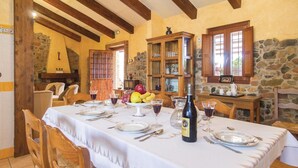 Un comedor con una mesa preparada para cuatro personas, con mantel blanco, copas de vino y una botella de vino. Se observan paredes de piedra y vigas de madera al fondo.