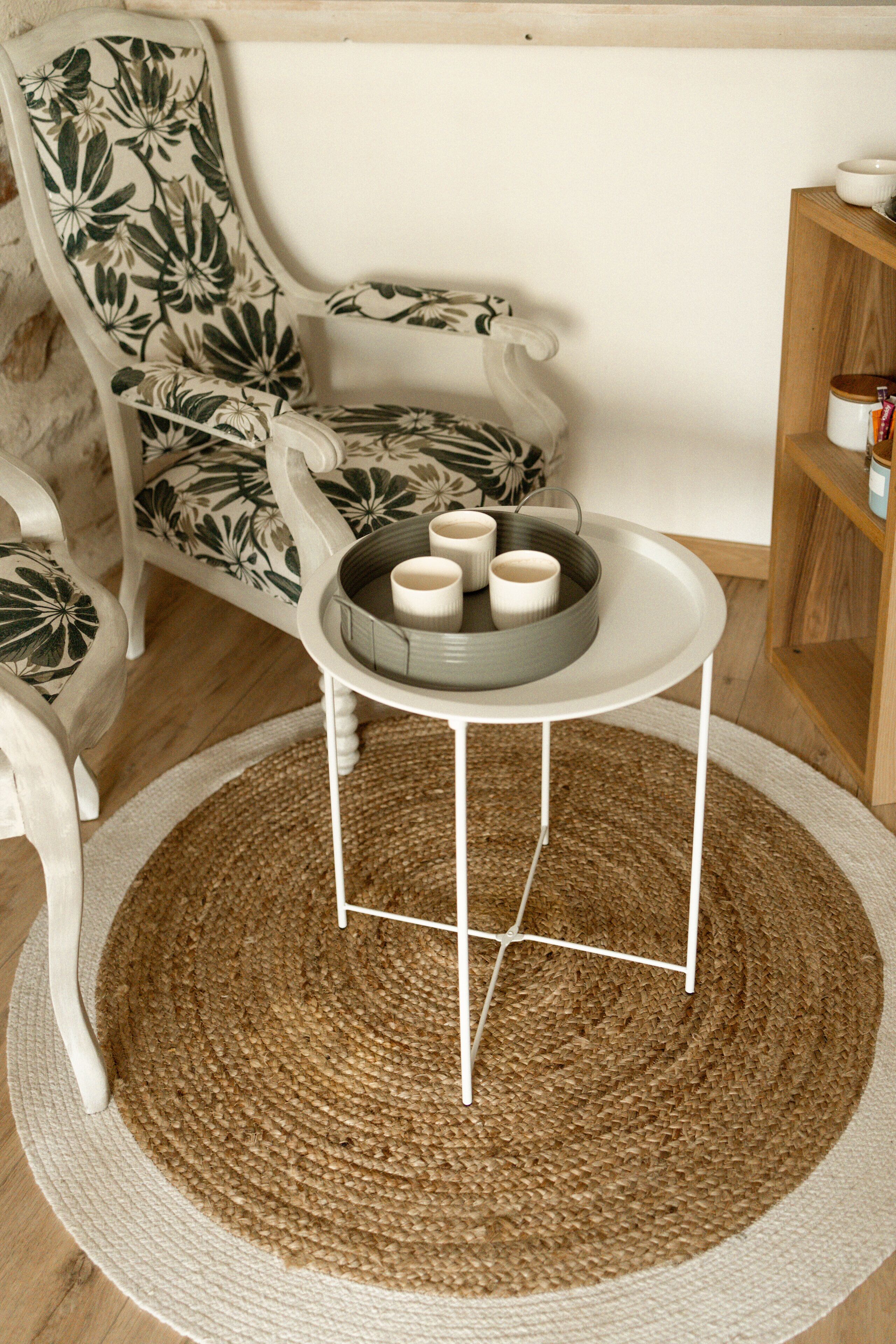 A round woven rug, a white chair with floral upholstery, and a small white table with a tray holding cups and a bowl.