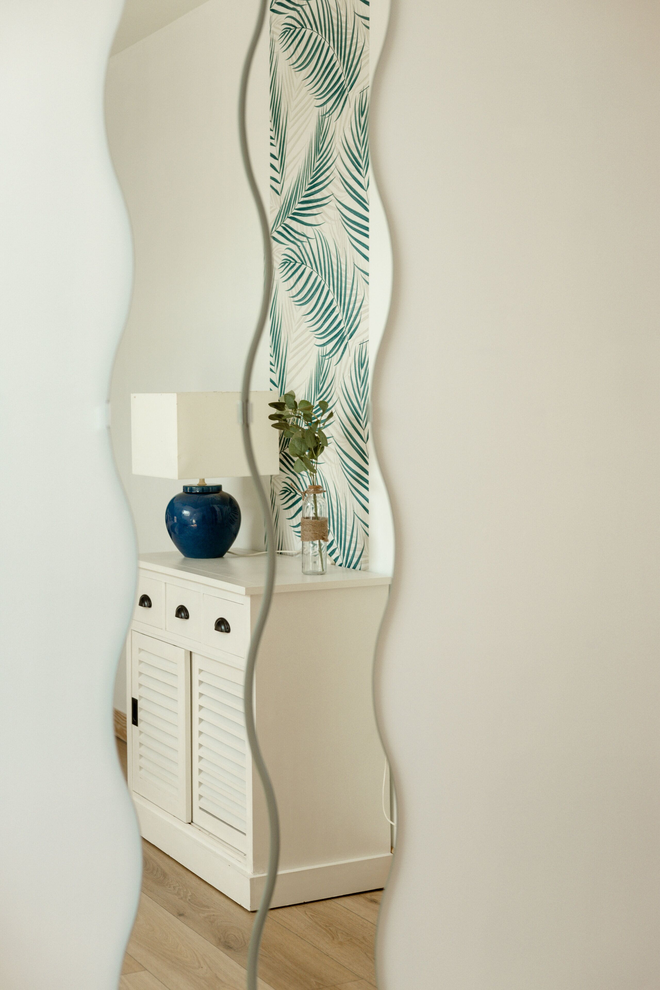 A room with a white cabinet, a blue vase, and a plant on a white wall with a leaf pattern.