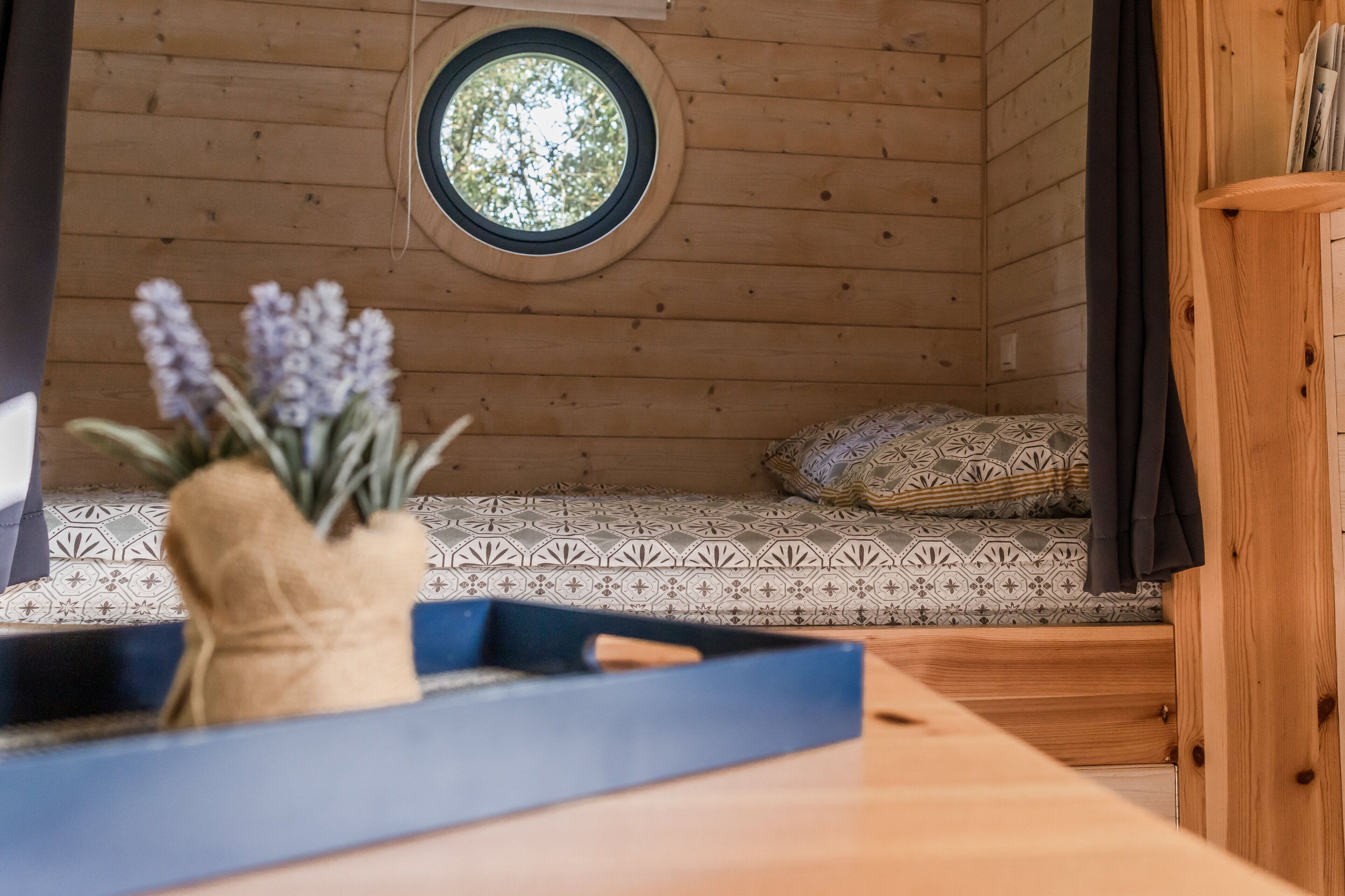 A cozy cabin interior with a wooden wall, a round window, a bed with patterned bedding, and a small table with a vase of flowers.