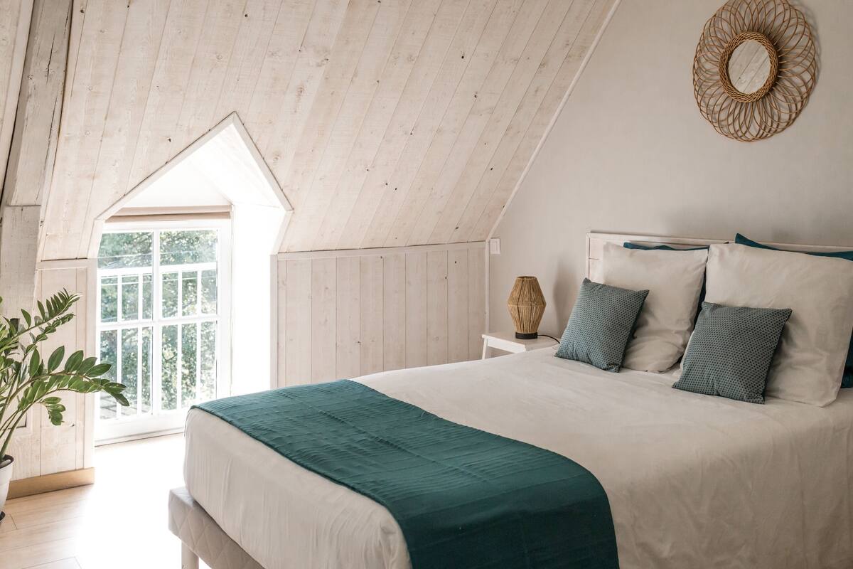 Un dormitorio con techo inclinado, una cama grande con una manta color turquesa, una lámpara de mimbre y vistas a través de una ventana.