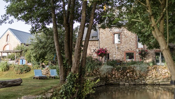 Un bâtiment en pierre avec un étang, entouré d’arbres et d’un petit jardin.