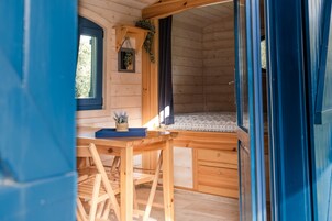 Interior de una cabaña de madera con comedor, una ventana pequeña y una cama con ropa de cama estampada.