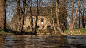 Un bâtiment en pierre doté de nombreuses fenêtres, entouré d’arbres et bordé par une rivière.