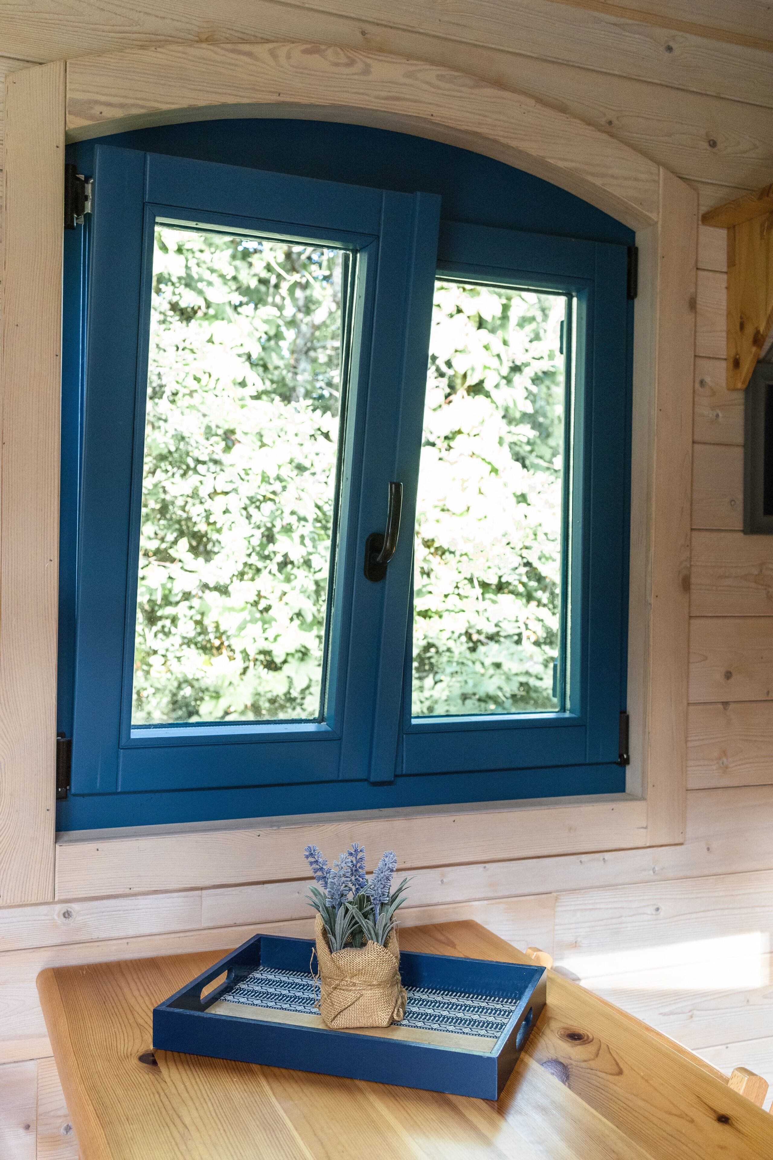 Een kamer met een houten interieur, een raam met blauwe lijst en een klein tafeltje met dienblad en een plant.