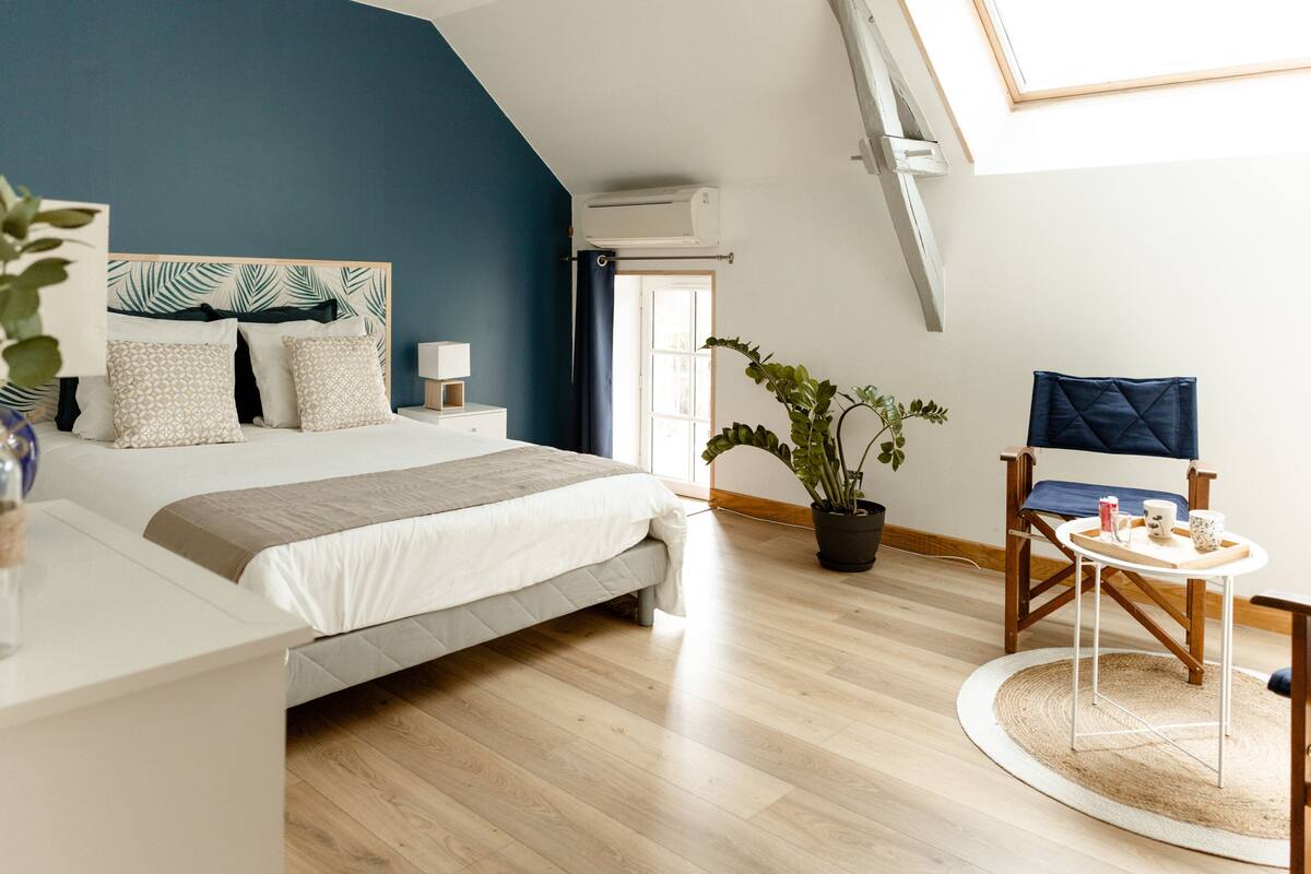 Un dormitorio con cama, mesita de noche, silla, planta en maceta y una pequeña mesa con bandeja.