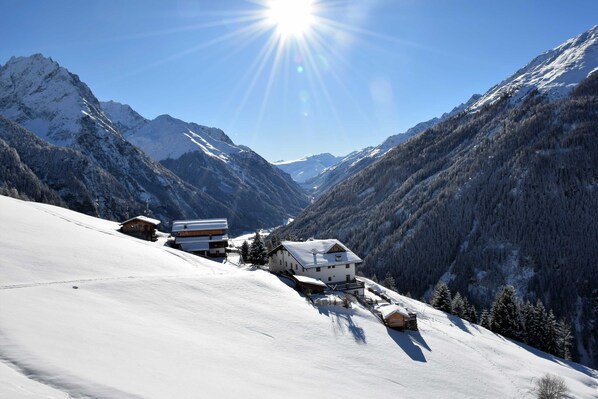 A snow-covered mountain landscape with a clear sky, a sun shining, and a few buildings at the base of the mountains.