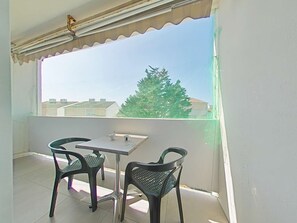 A balcony with a table and two chairs, a view of a tree and buildings, and a window with a curtain.