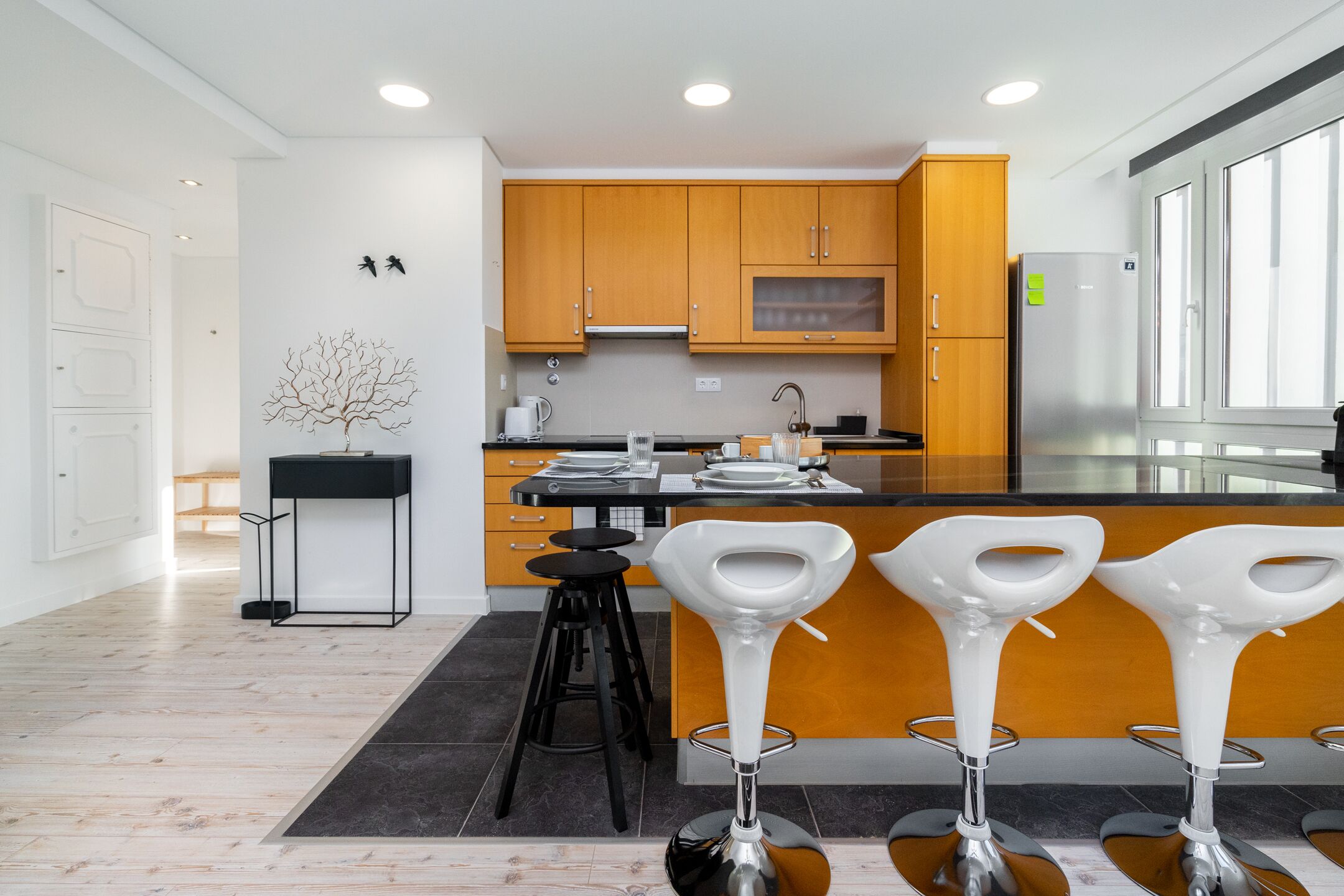 A modern kitchen with wooden cabinets, a stainless steel refrigerator, and a bar area with white stools.
