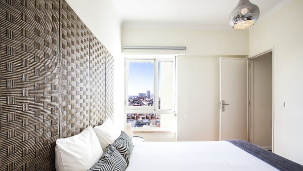 Un dormitorio con un cabecero estampado, una cama con ropa de cama blanca y a rayas, vista de la ciudad a través de una ventana abierta y una lámpara colgante.