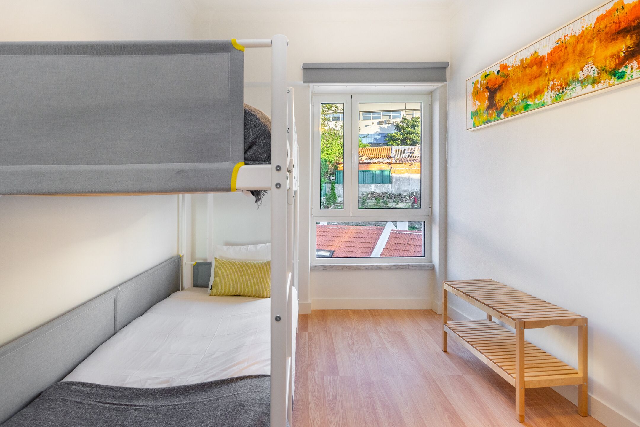 A room with a bunk bed, a small wooden bench, and a window with a view of buildings.