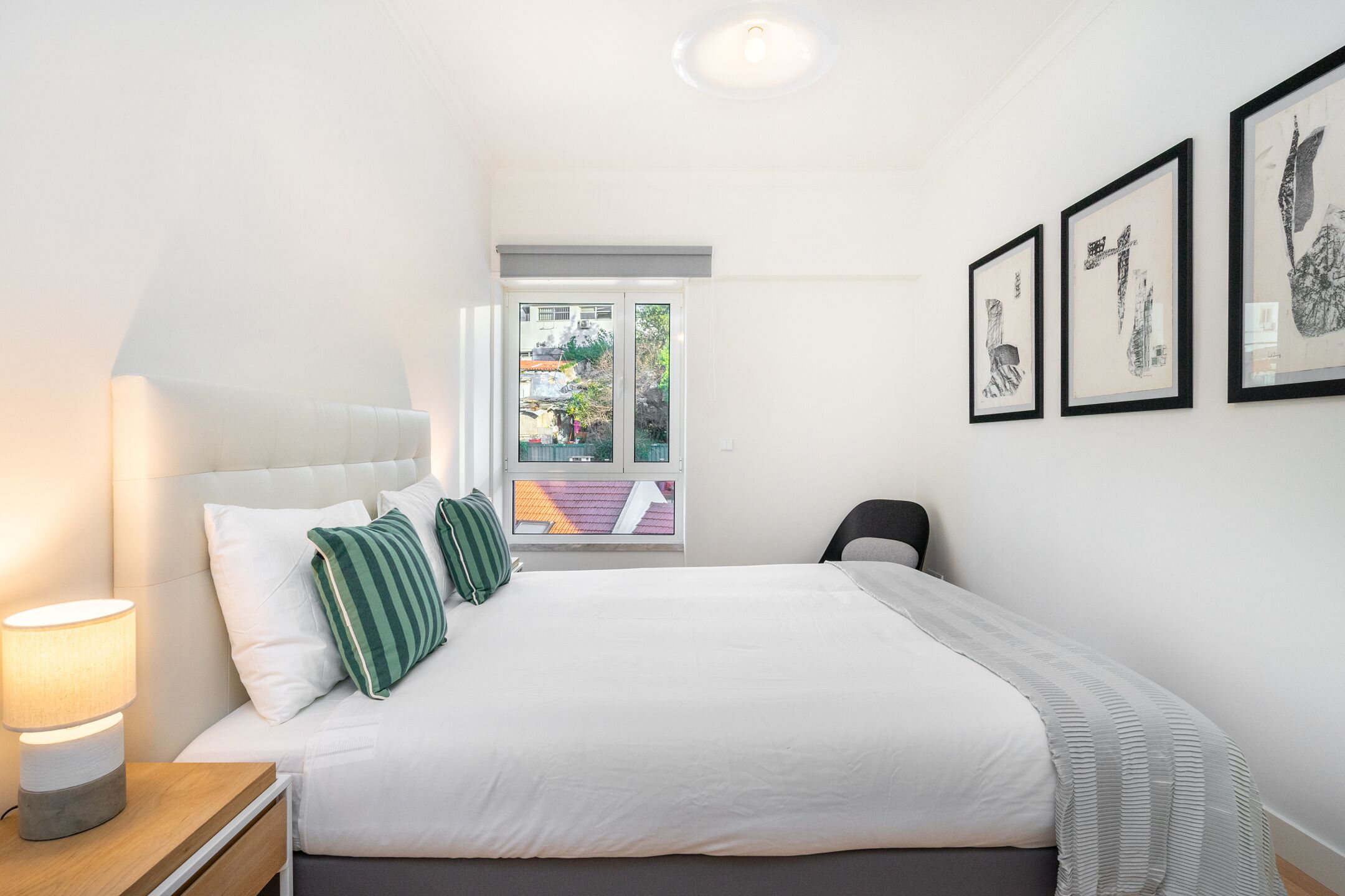 Une chambre à coucher avec un lit, une table de chevet, une lampe, une fenêtre et des œuvres d’art encadrées accrochées au mur.
