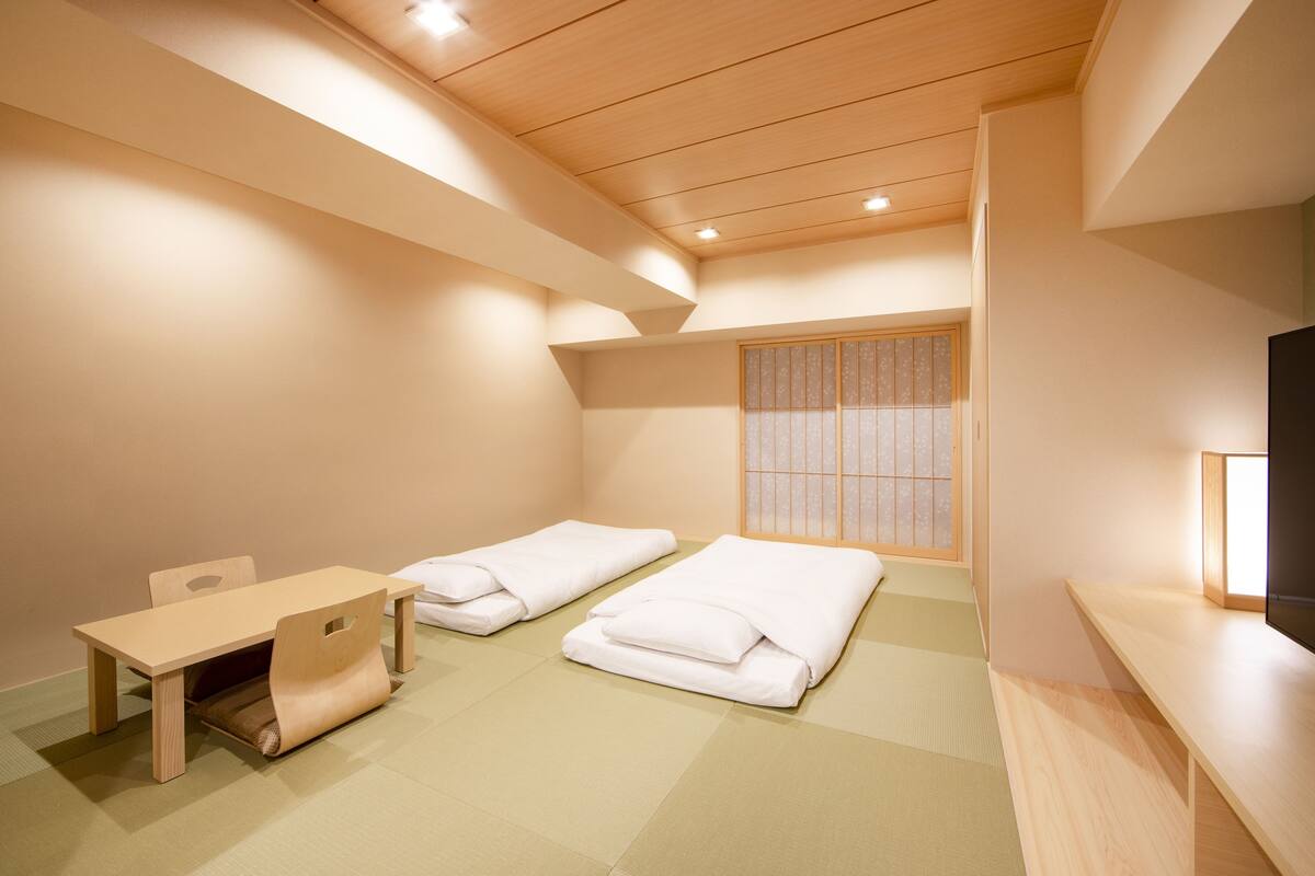A room with a low ceiling, tatami mat flooring, a wooden desk, two beds with white bedding, and a TV on a wooden stand.