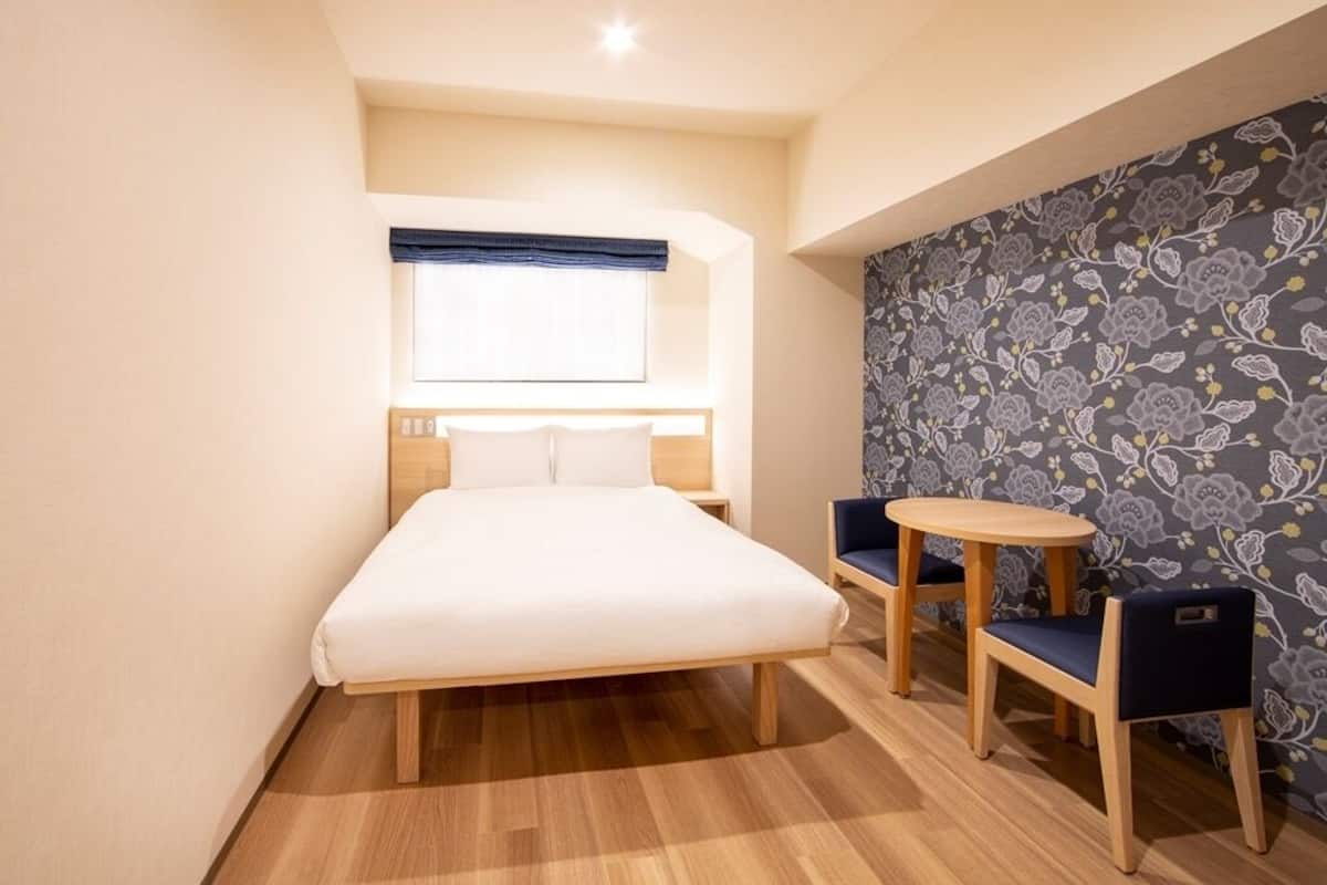 A bedroom with a bed, a small round table, two chairs, a window with blinds, and a patterned wallpaper.