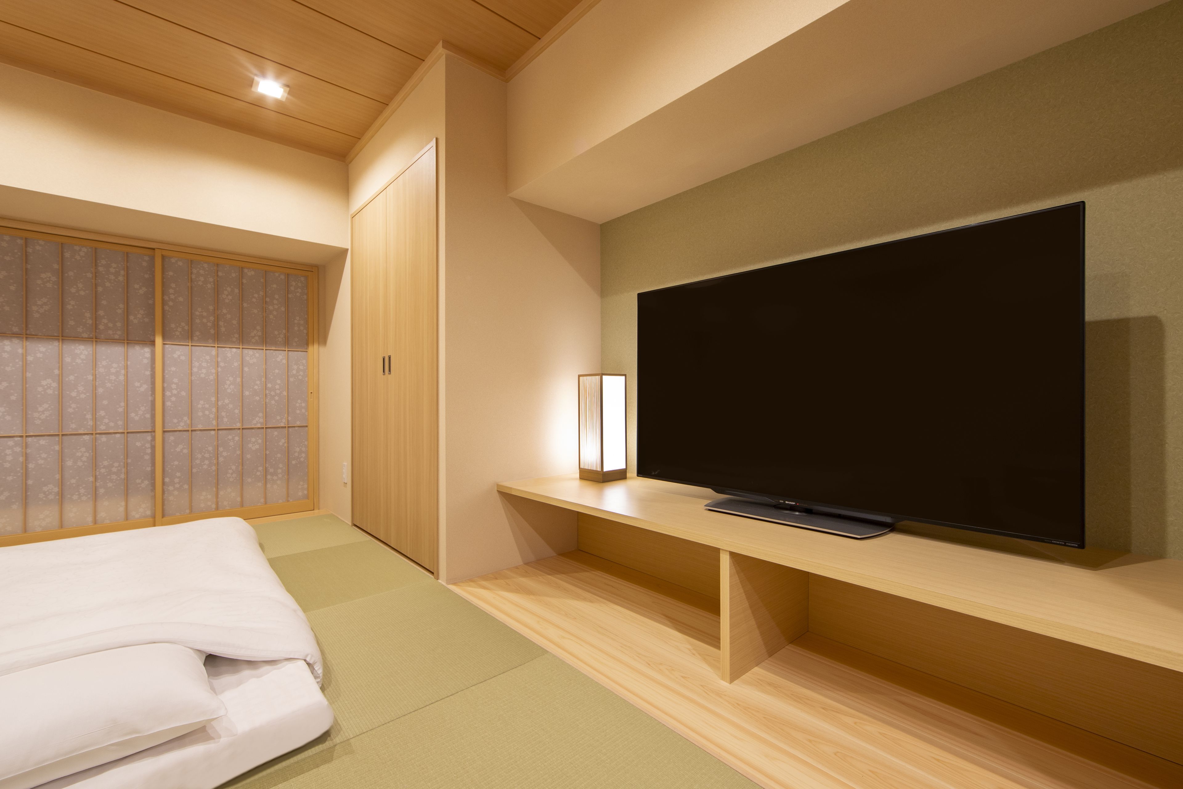 A modern hotel room with a large flat-screen TV on a wooden stand, a bed with white bedding, a traditional sliding door, and a small bedside table with a lamp.