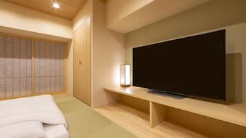 A modern hotel room with a large flat-screen TV on a wooden stand, a bed with white bedding, a traditional sliding door, and a small bedside table with a lamp.