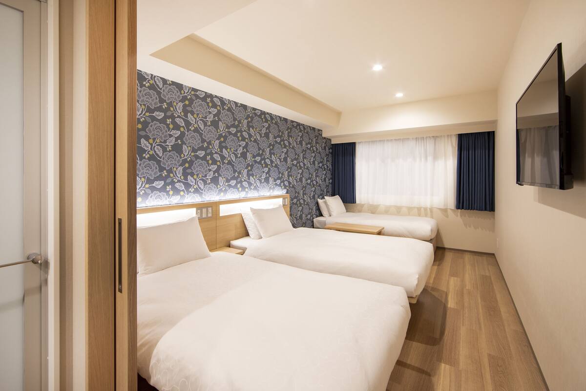 A hotel room with two beds, a large window with curtains, a flat-screen TV, and a floral-patterned wall.