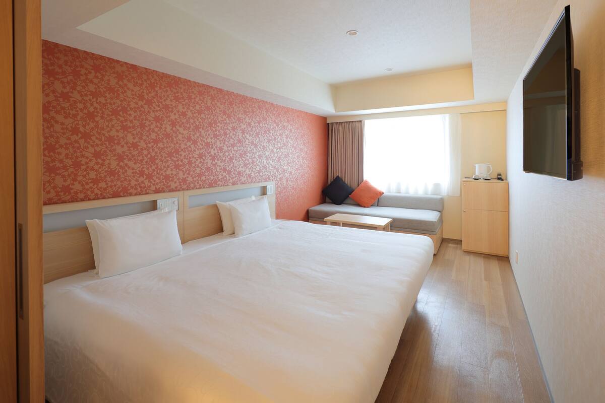 A hotel room with a large bed, a desk, a chair, a TV, and a wardrobe.