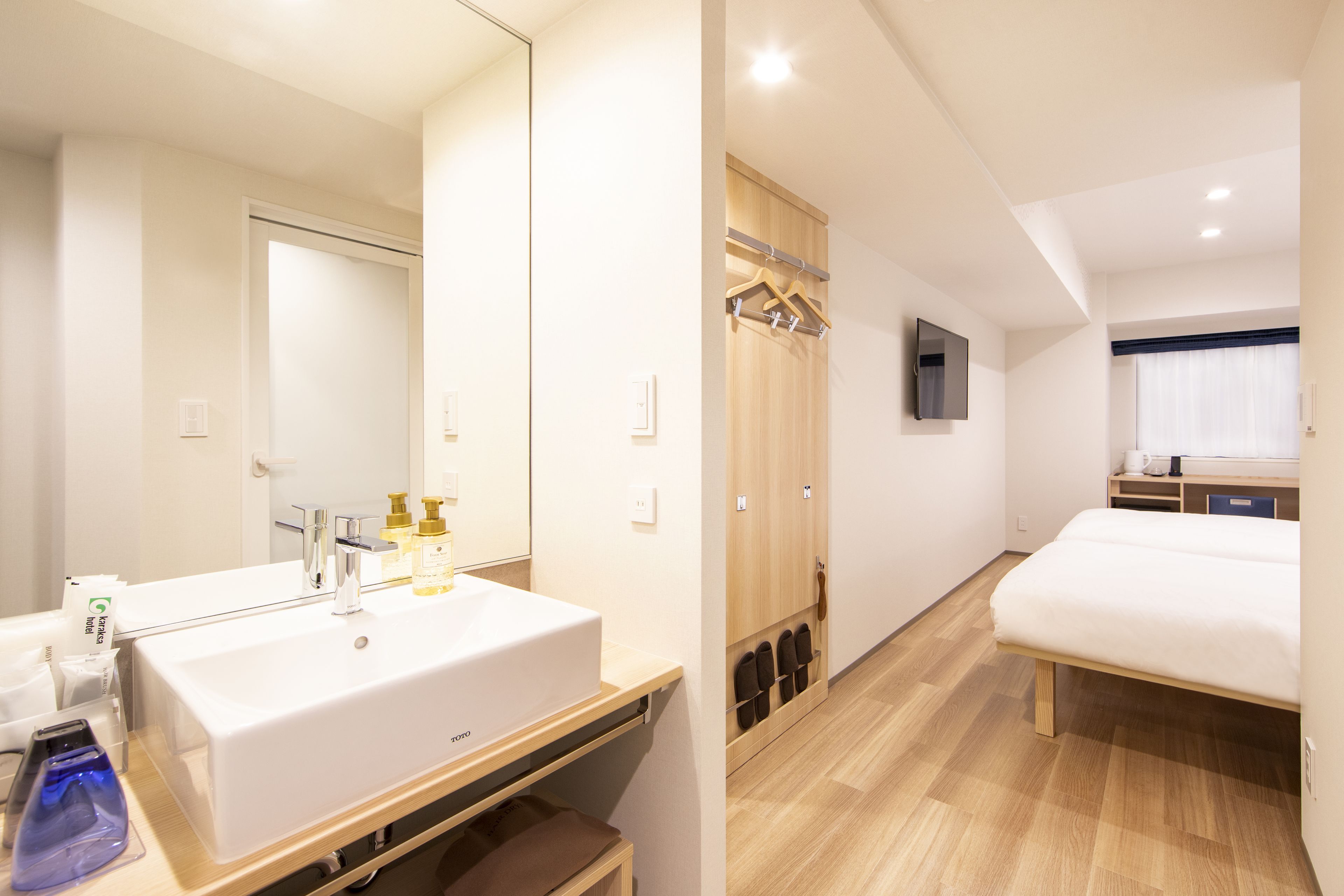 A hotel room with a large white sink, a mirror, a wooden wardrobe, a TV mounted on the wall, and a bed.
