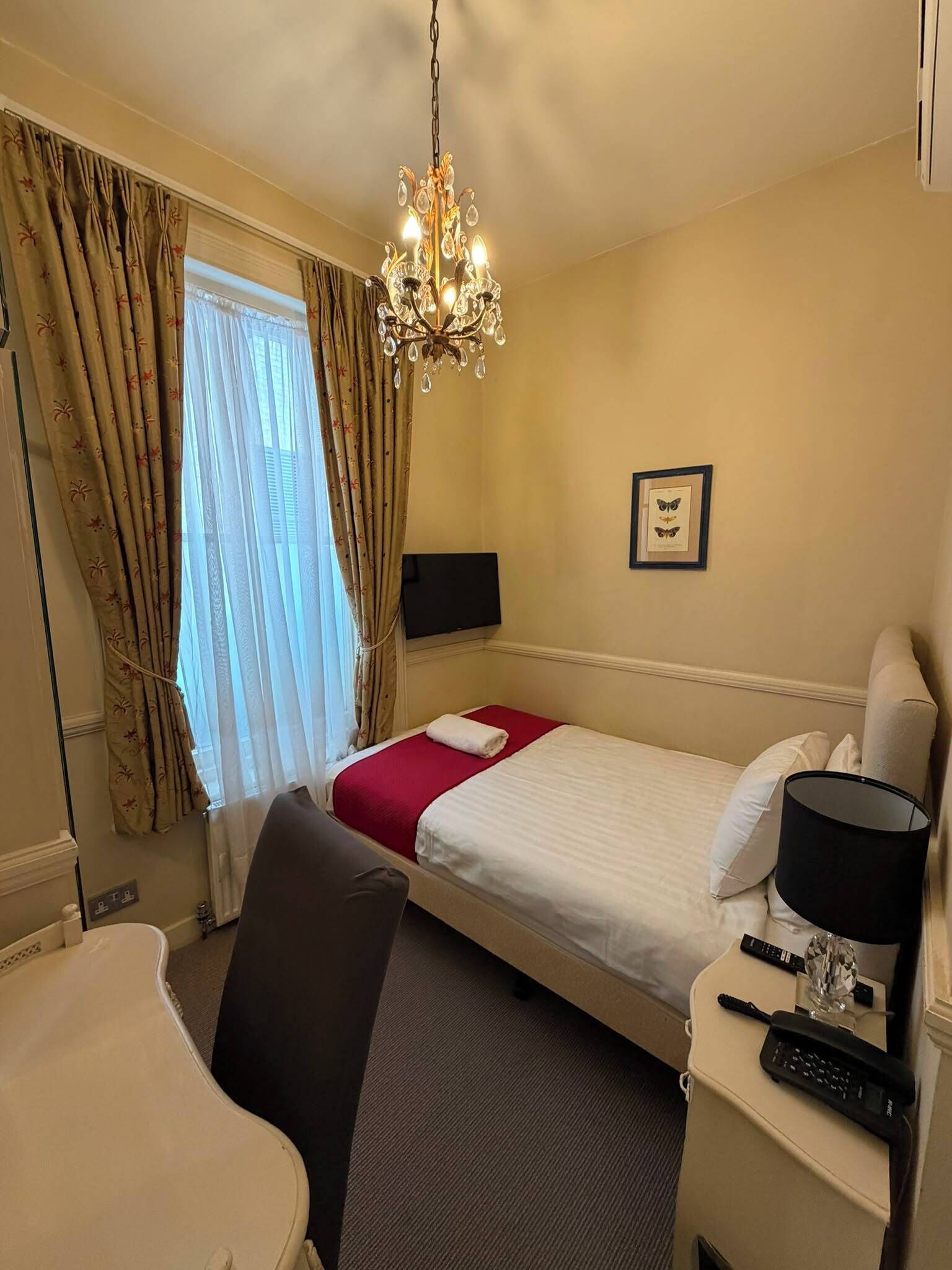 A hotel room with a bed, a desk, a chair, a telephone, a lamp, a chandelier, and a window with curtains.