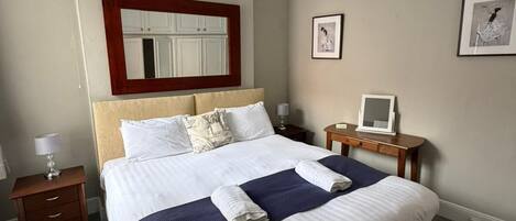 A neatly made bed with a headboard, a bedside table with a lamp, a small bedside table with a mirror, and a framed picture on the wall.