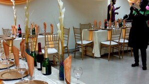 Un salón de banquetes con mesas dispuestas para un evento formal, destacando manteles blancos, servilletas doradas y centros de mesa florales.