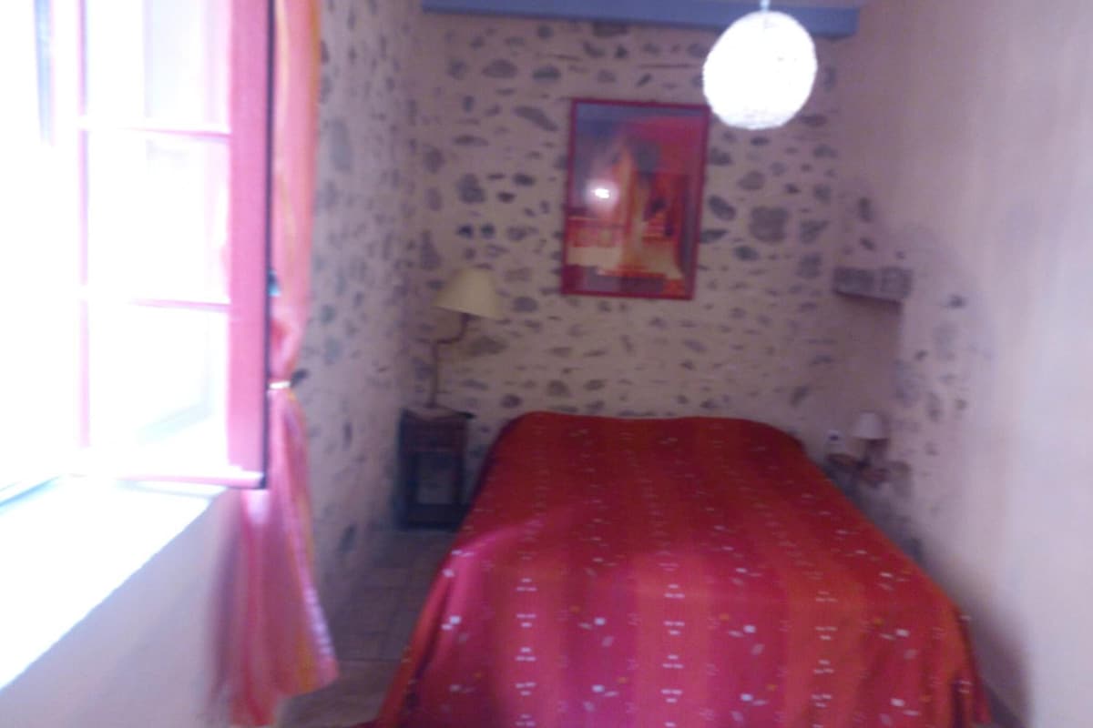Una habitación estrecha con una cama, un ventanal con cortinas rosadas, una lámpara y una pared con un cartel.